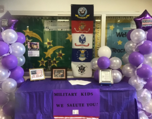 Military flags; purple and white balloon on table covered with purple tablecloth. Message "Military Kids We Salute you." 