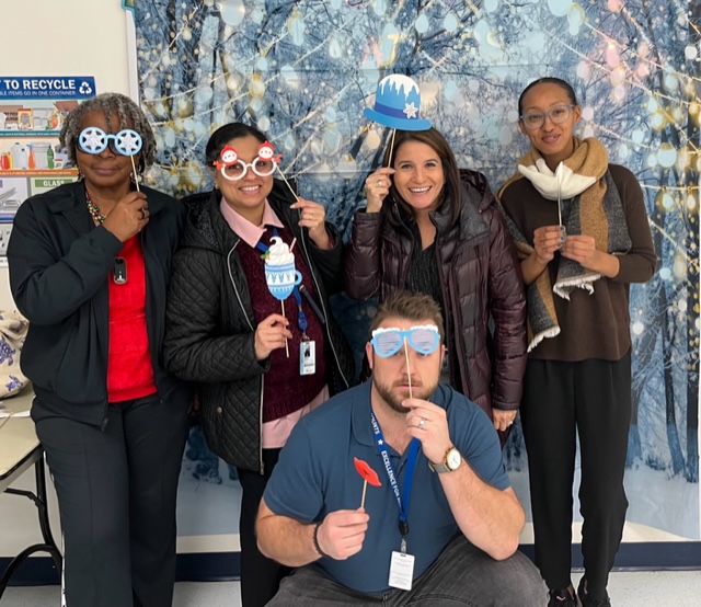 Teachers at a holiday theme photo booth (each holding different snow flakes). A male teacher squad down and four female teachers standing behind.