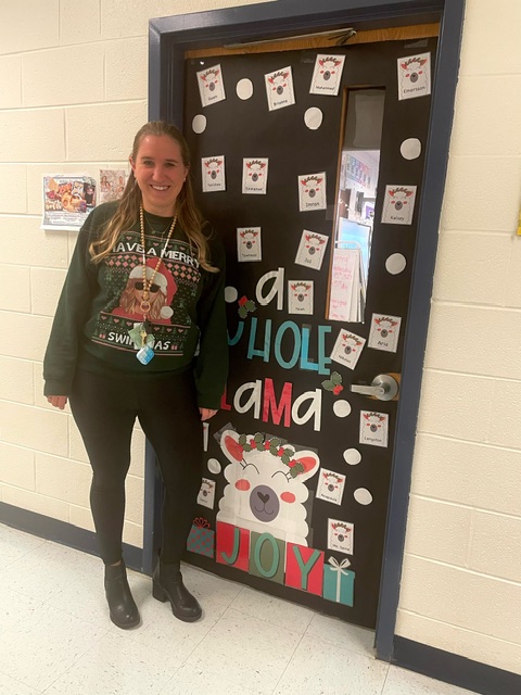 kindergarten teacher wearing Santa sweater and stand next to a door has holiday decoration.