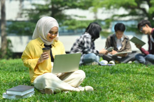 summer digital learning; girl siting on grass with laptop; 3 students sitting in a group in the background.