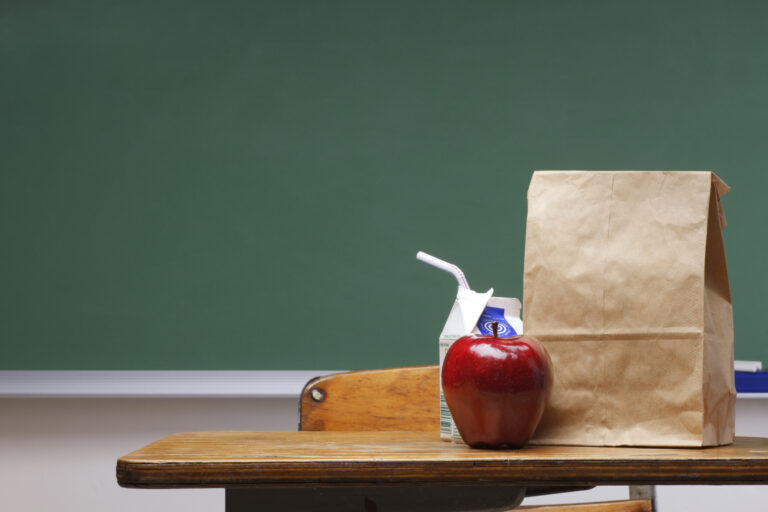 Student desk; red apple, milk carton and brown lunch bag on desk.